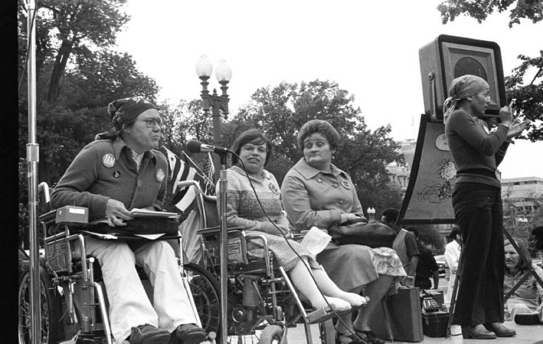 Black and white photograph showing a group of disability rights activists, including women in wheelchairs gathered outdoors at a rally. The activists wear determined expressions as they advocate for accessibility and equal rights.