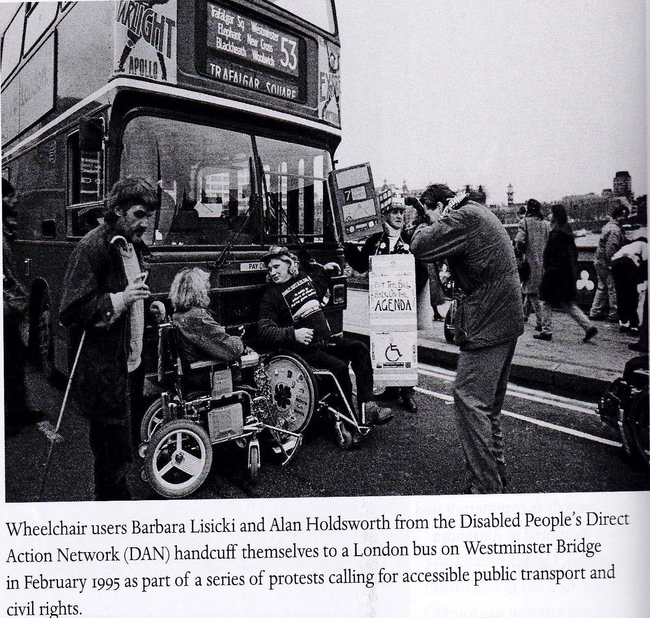 Black and white photo shows wheelchair users Barbara Lisicki and Alan Holdsworth handcuffed to a London bus on Westminster Bridge in February 1995. Activists from the Disabled People's Direct Action Network (DAN) surround the bus, protesting for accessible public transport and civil rights.