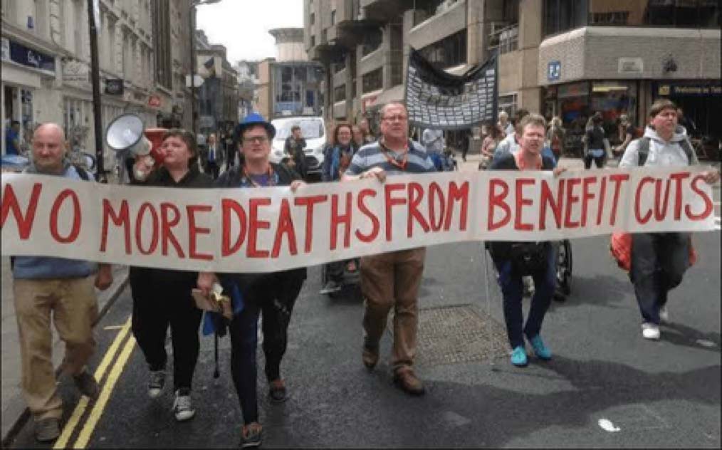A group of people walk down a city street holding a long banner that reads "NO MORE DEATHS FROM BENEFIT CUTS" in red block letters.