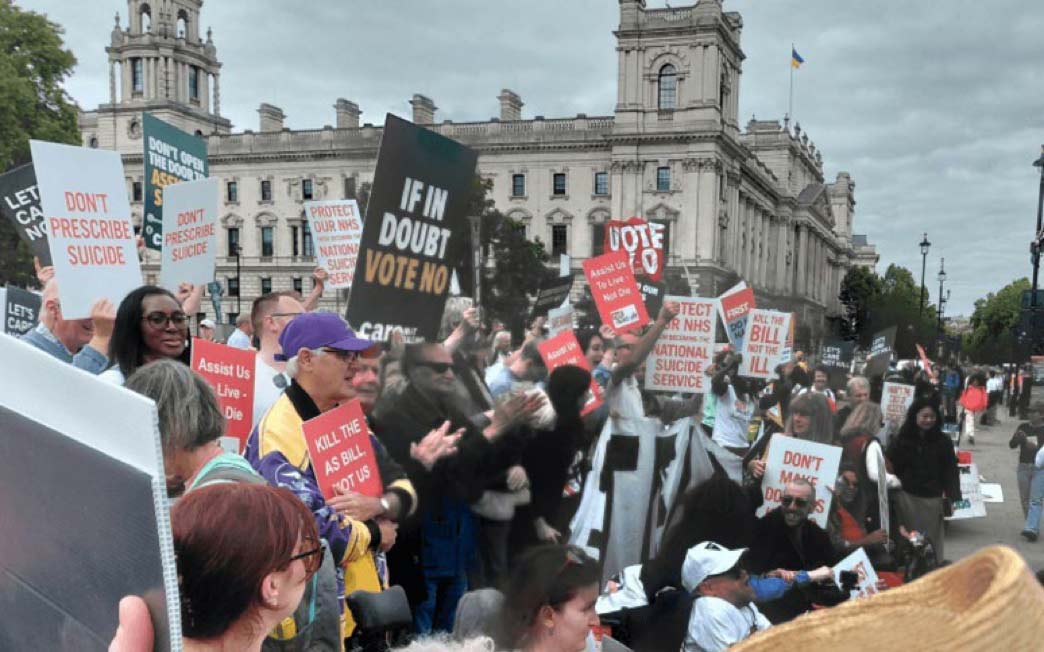 A group of protestors gather outside a large, classical stone government building in London, holding signs with anti-euthanasia and anti-assisted dying messages such as 'Don't Prescribe Suicide', 'If In Doubt Vote No', and 'Kill The Bill Not Us'. The atmosphere is tense and focused, with diverse participants expressing opposition to assisted dying legislation.